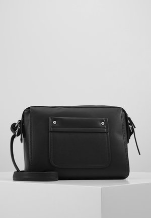 Bolso bandolera rectangular negro con bolsillo frontal de solapa, remaches plateados y correa ajustable para el hombro sobre una superficie blanca y fondo gris claro.