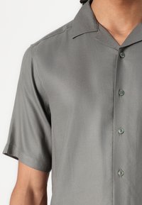 Short-sleeved, collared shirt in a muted gray hue, made of smooth fabric with six light green buttons and clean stitching details.