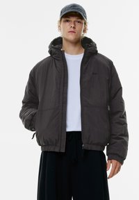 Zwarte hooded jacket van lichtgewicht materiaal, met een rits aan de voorkant, elastische manchetten en zijzakken. Gemaakt over een wit shirt.