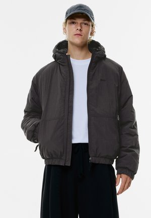 Black hooded jacket made of lightweight material, featuring a front zipper, elastic cuffs, and side pockets. Worn over a white shirt.