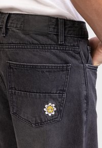 Un jean en denim noir avec une poche arrière ornée d'un patch représentant un tournesol souriant. Le matériau est robuste avec une finition texturée.