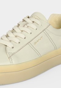 Baskets blanches en cuir avec semelle beige, lacets blancs et logo doré "GANT" sur le côté et la languette.