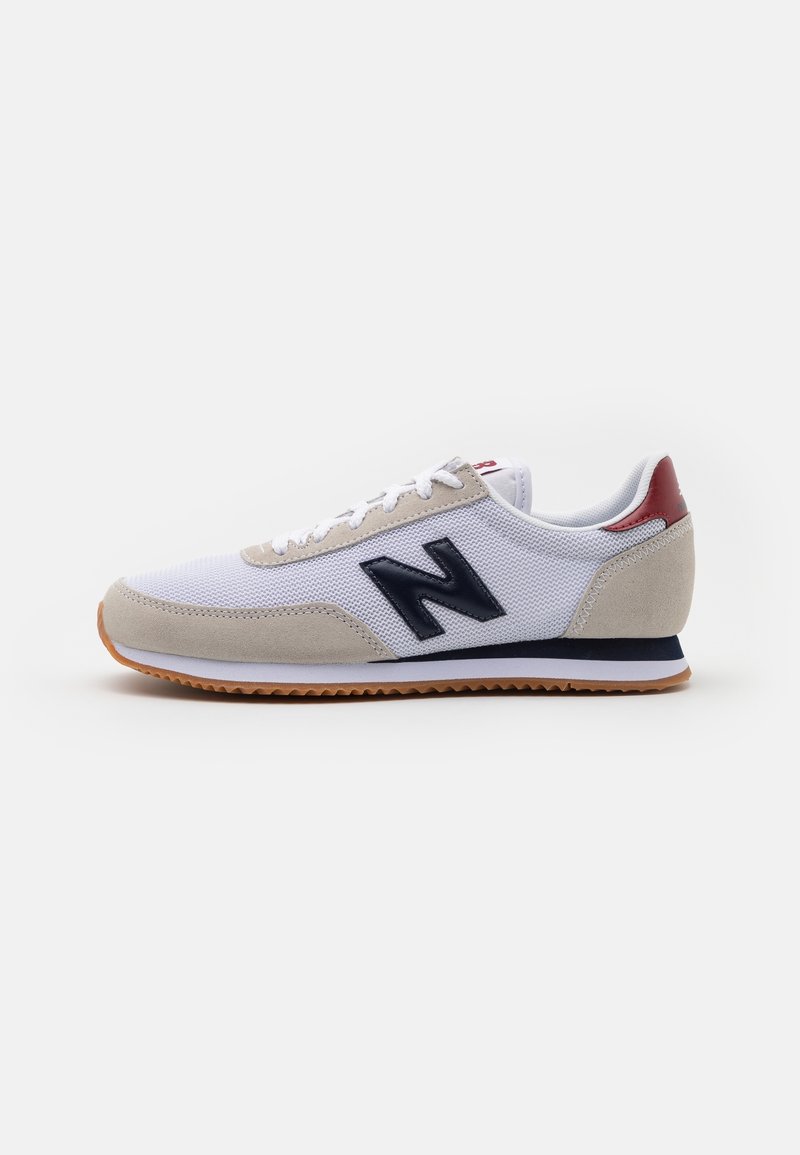 Chaussure de sport blanche et beige avec logo "N" noir, semelle en caoutchouc naturel, et accent rouge au talon, affichée de côté intérieur sur un fond blanc.