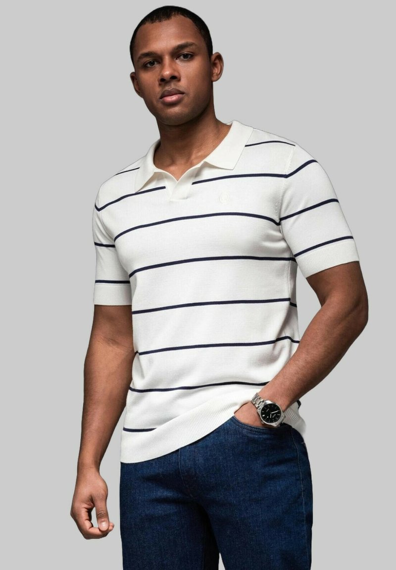 Vit kortärmad polo med marinblå ränder, ribbad krage och ärmslut, tillsammans med mörkblå jeans; modellen bär en silverfärgad klocka på handleden.