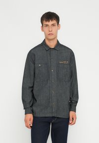 Camicia button-up grigia scura con un motivo a righe, due tasche sul petto e ricami dorati. La trama del tessuto appare liscia.
