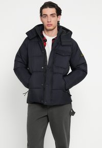 Granatowa pikowana kurtka puchowa z odpinanym kapturem, zamkiem błyskawicznym, zatrzaskami, wieloma kieszeniami i ściągaczem u dołu. Gładka, pikowana faktura.