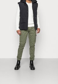 Svart vadderad väst över en vit kabelstickad tröja, olivgröna cargobyxor och svarta ankelskor. Slät textur med funktionella fickor.