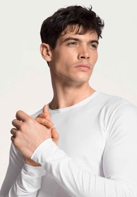 Long-sleeve white top with a round neckline, smooth fabric, fitted design, and no visible patterns. Light skin tones and short, dark hair.