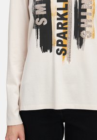 Jasnobeżowa koszula z długim rękawem, z czarno-złotym wzorem przypominającym pociągnięcia pędzla, zawierająca tekst i akcenty z kryształków. Fason o usztywnionej konstrukcji.