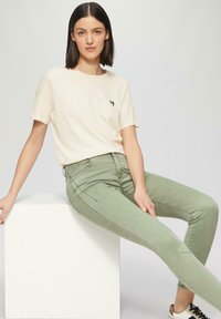 Jasno beżowa koszulka z krótkim rękawem z małym czarnym logo, w po�łączeniu z dopasowanymi jasnooliwkowymi dżinsami i casualowymi trampkami.