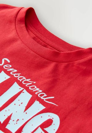 Camiseta de algodón roja con cuello redondo, con texto gráfico blanco que dice "Sensation" y "KING" en una fuente audaz y divertida.