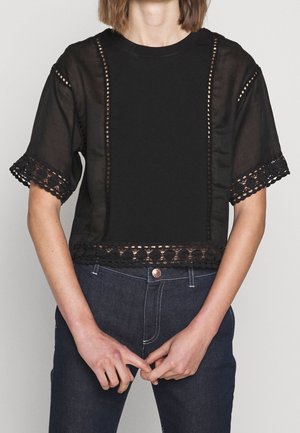 Person iført sort kortærmet bluse med blonde detaljer på ærmer og kant, kombineret med mørkeblå jeans, hænder foldet foran.