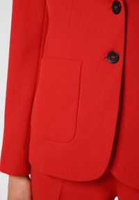Veste de costume rouge en tissu lisse, coupe ajustée, simple boutonnage avec deux boutons noirs et détail de poche plate.