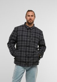 Melna un pelēka kvadrātniecveida jaka ar apkakli, pogu aizdari un sānu kabatām. Audums izskatās mīksts ar teksturētu apdari.