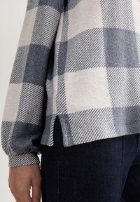 Langsleeve top in grijs en witte ruitpatronen, gemaakt van zachte stof met een lichte zijsplit, gecombineerd met donkere spijkerbroek.