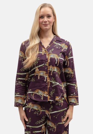 Vrouw draagt een bijpassende donkerpaarse pyjama set met een luipaard- en blaadjestakpatroon, staand tegen een eenvoudige lichte achtergrond.