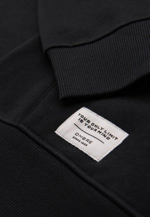 Schwarzer Sweatshirt mit gerippten Bündchen und Saum. Enthält ein weißes Stoffetikett mit dem Text: "DEIN EINZIGES LIMIT IST DEIN GEIST" und "OMBRE seit 1985."