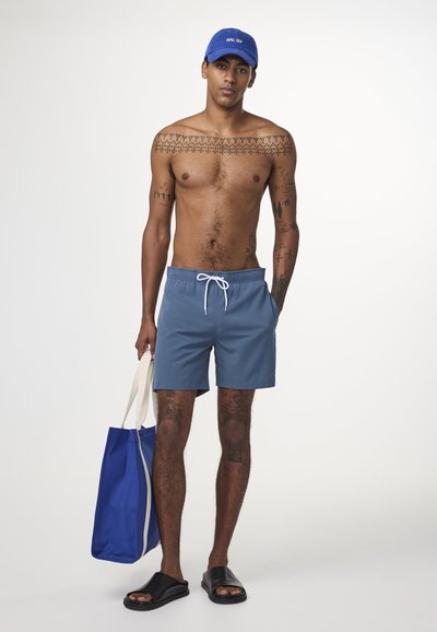 Shorts de bain bleus avec cordon blanc, sandales noires, casquette bleue avec écriture blanche et sac fourre-tout bleu. Des tatouages couvrent le haut du corps et les jambes.