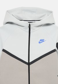 Lys grå hettegenser med svart og beige aksenter, med et chevron-design, hette og en blå Nike-logo på brystet.