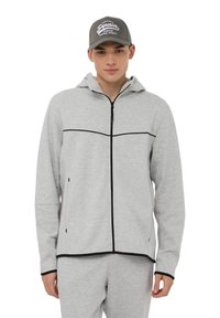 Felpa grigia con zip e cappuccio, caratterizzata da accenti orizzontali neri sul petto e tasche laterali. Indossata con pantaloni grigi coordinati e un cappellino.