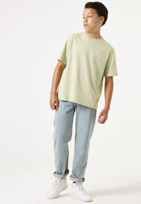 Camiseta de algodón verde claro con mangas cortas y un pequeño gráfico en el lado izquierdo del pecho, combinada con unos pantalones vaqueros azul claro de corte holgado y zapatillas blancas.