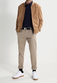Tan suède bomberjack over een marineblauw polo, gecombineerd met beige broeken. Witte sneakers met bruine zolen. Eenvoudige, casual outfit met opgerolde mouwen.