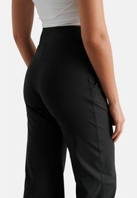 Pantaloni neri a vita alta con una texture liscia, dotati di tasche laterali e dettagli di cucitura, visti da dietro.