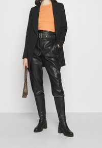 Blazer nero a righe sottili sopra una maglietta arancione, abbinato a pantaloni in finta pelle nera a vita alta e stivali neri al ginocchio con tacco.