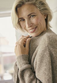 Femme souriante avec les cheveux blonds courts ajustant le col de son pull en tricot beige près d'une fenêtre lumineuse.
