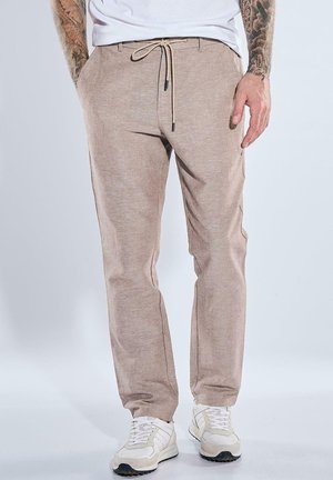 Mann trägt beige Kordelzughose, weiße Sneakers, weißes T-Shirt, mit tätowierten Unterarmen sichtbar.