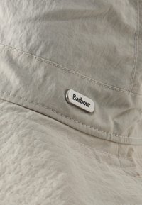 Καπέλο τύπου bucket από απαλή μπεζ ύφανση με ραμμένα ραφές και μικρή μεταλλική ετικέτα με την ένδειξη "Barbour" στο πλάι.