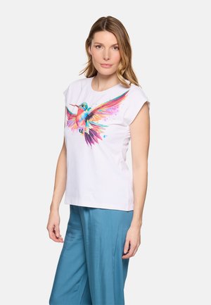 Mujer con cabello castaño claro, vistiendo una blusa blanca sin mangas con estampado colorido de colibrí y pantalones azules sueltos, de pie contra un fondo blanco.