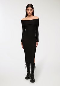 Abito nero a costine con scollo a spalle scoperte, maniche lunghe e spacco laterale. Abbinato a stivali neri alti fino al ginocchio con lacci.