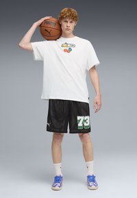 Camiseta blanca de manga corta con gráfico, pantalones cortos de baloncesto de malla negra con el número verde "73", y zapatillas azules con patrón. Sosteniendo una pelota de baloncesto marrón.