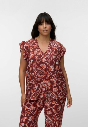 Kvinde iført en rød blomstret kortærmet bluse og matchende bukser, stående mod en ensartet lys baggrund.