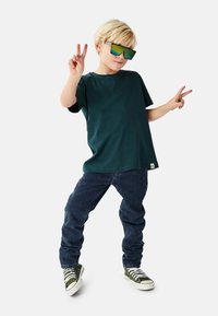 Grøn bomuld T-shirt, løs pasform, kombineret med mørkeblå denimjeans. Barnet bærer store reflekterende solbriller og grønne/hvide sneakers.