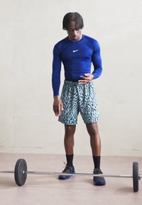 Blaues Langarm-Sportoberteil, gemusterte aquafarbene Shorts, schwarze Schuhe, steht neben einer Langhantel auf einem hellen Boden, neutraler Hintergrund.
