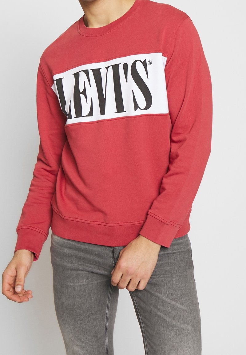 Červená mikina s bílým pruhem, na kterém je černé logo "LEVI'S". Design s kulatým výstřihem, měkký materiál, žebrované manžety a lem. Spárováno se šedými džíny.