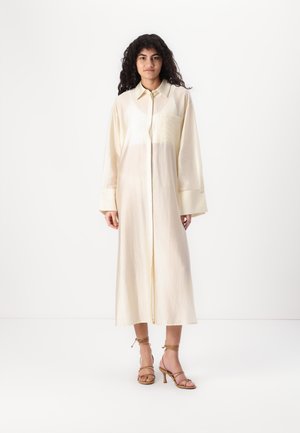Rochie lungă, de culoare crem, cu butoni, realizată dintr-un material ușor, având o buzunar frontal și mâneci largi. Purtați cu sandale bej cu curelușe.