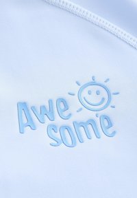 Heledat sinine kangas, millel on tikitud tekst "Awe some" koos naeratava päikese ikooniga. Pehme tekstuur ja puhas, modernne disain.