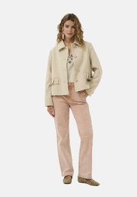 bézs gyapjú zakó masnis díszítésű zsebekkel és kerek gombokkal, világos rózsaszín kordbársony nadrággal és dekoratív lapos cipőkkel párosítva.