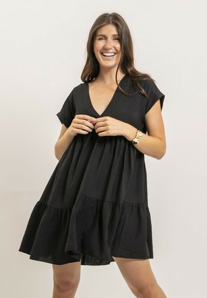 Femme souriante aux longs cheveux bruns portant une robe noire ample à manches courtes et un bracelet en or, debout devant un fond clair uni.