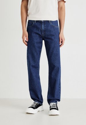 Person, der dunkelblaue gerade geschnittene Jeans, ein weißes T-Shirt und schwarze und weiße Turnschuhe trägt, steht auf einem hellen Boden vor einem schlichten Hintergrund.