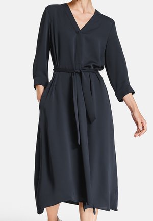 Kobieta ubrana w czarną, midi sukienkę z długim rękawem, dekoltem w serek oraz paskiem związanym w talii, pozująca z rękami częściowo wsuniętymi do kieszeni.