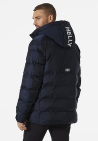 Chaqueta acolchada navy con capucha, acolchado visible y logo de Helly en la parte posterior. El material parece suave y resistente al agua.