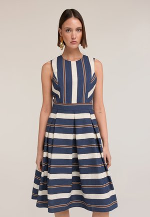 Femme portant une robe sans manches rayée bleu marine et blanc avec une jupe plissée, debout sur un fond beige uni, portant des boucles d'oreilles en or.