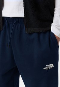 Pantalones de chándal azul marino con cintura elástica y cordón; presenta el logo blanco de The North Face en el muslo izquierdo. Material suave.