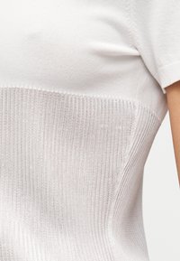 Κοντινή λήψη λευκού κοντομάνικου πλεκτού τoπ που δείχνει ένα λείο πάνω μέρος και ένα ριμπ κάτω μέρος στον κορμό ενός ατόμου.