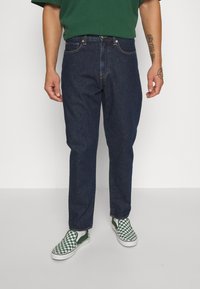 Blå denimjeans med rak skärning, fem fickor och guldtråd; bärs med grön- och vitrutiga slip-on skor.