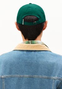 Person, der mit dem Rücken zur Kamera steht, trägt eine grüne Baseballmütze, eine Jeansjacke mit beigem Kragen und ein grün kariertes Hemd darunter, vor einem weißen Hintergrund.